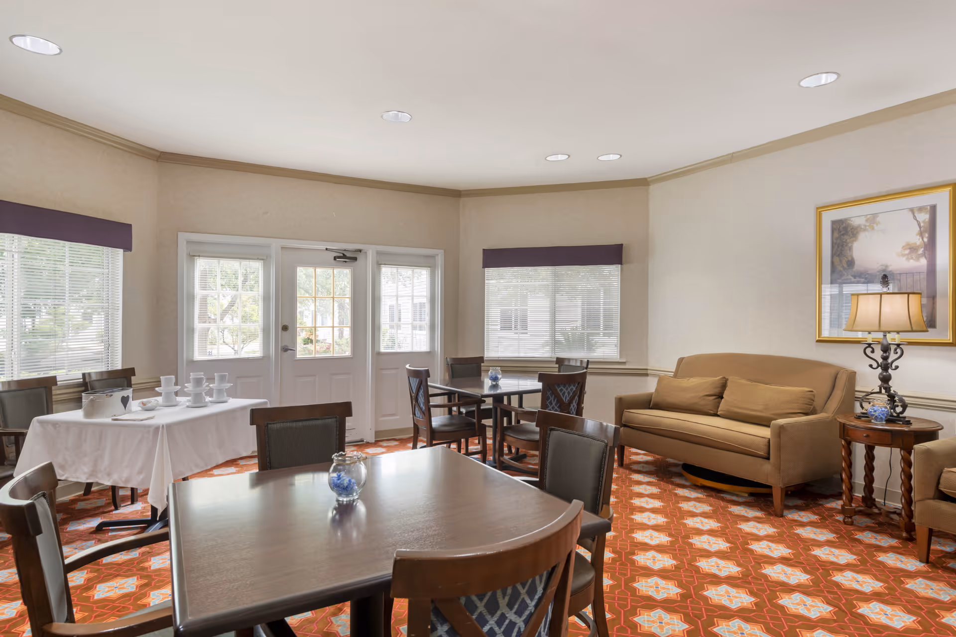 A bright and cozy common area in a senior living facility featuring multiple tables and chairs, a beige sofa with cushions, a side table with a lamp, large windows with blinds, and a door leading outside. The floor has a patterned carpet in shades of red and orange, and a framed picture hangs on the wall.