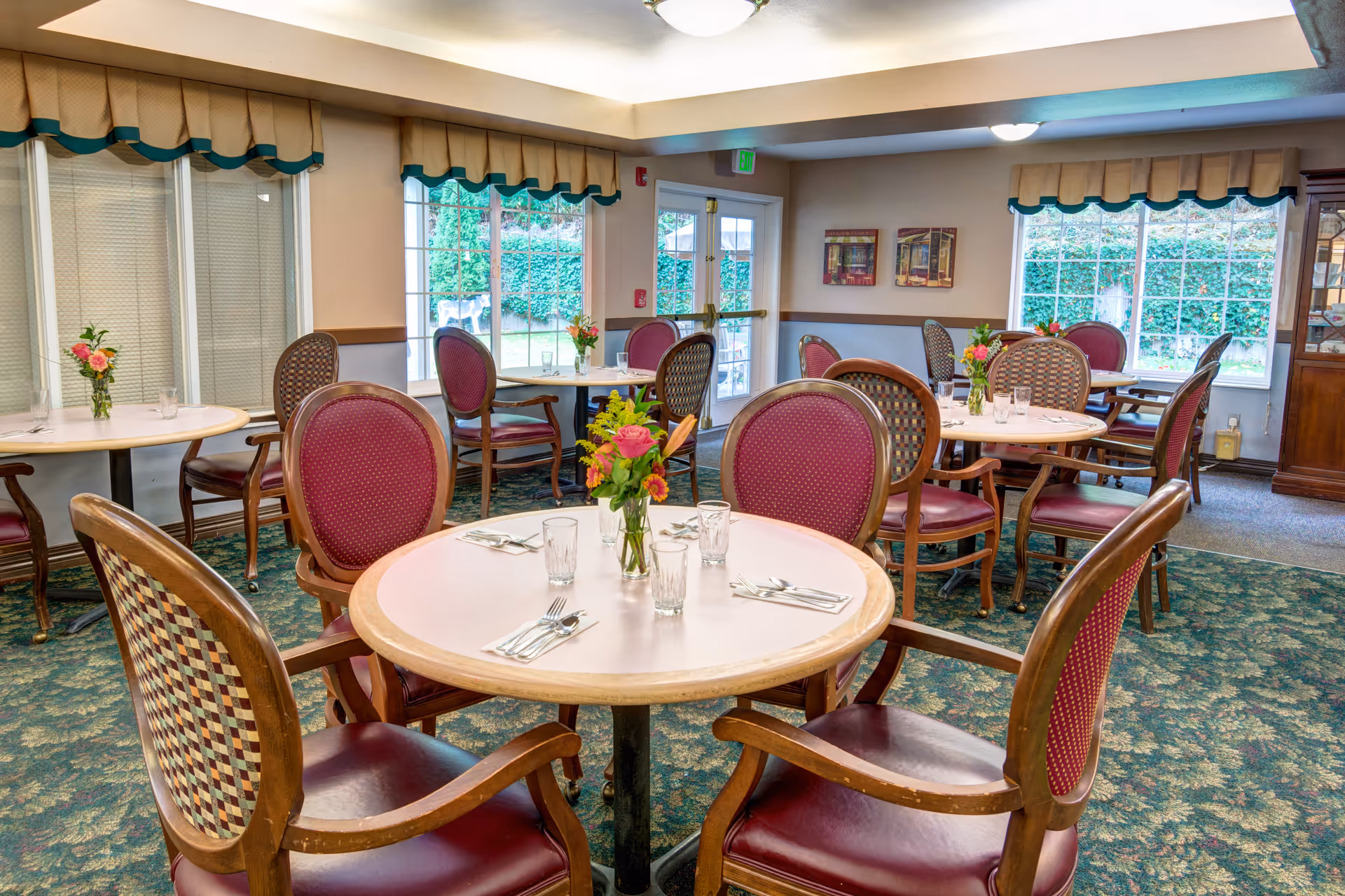 Bright dining room with round tables set with flowers, place settings, and upholstered chairs by large windows.