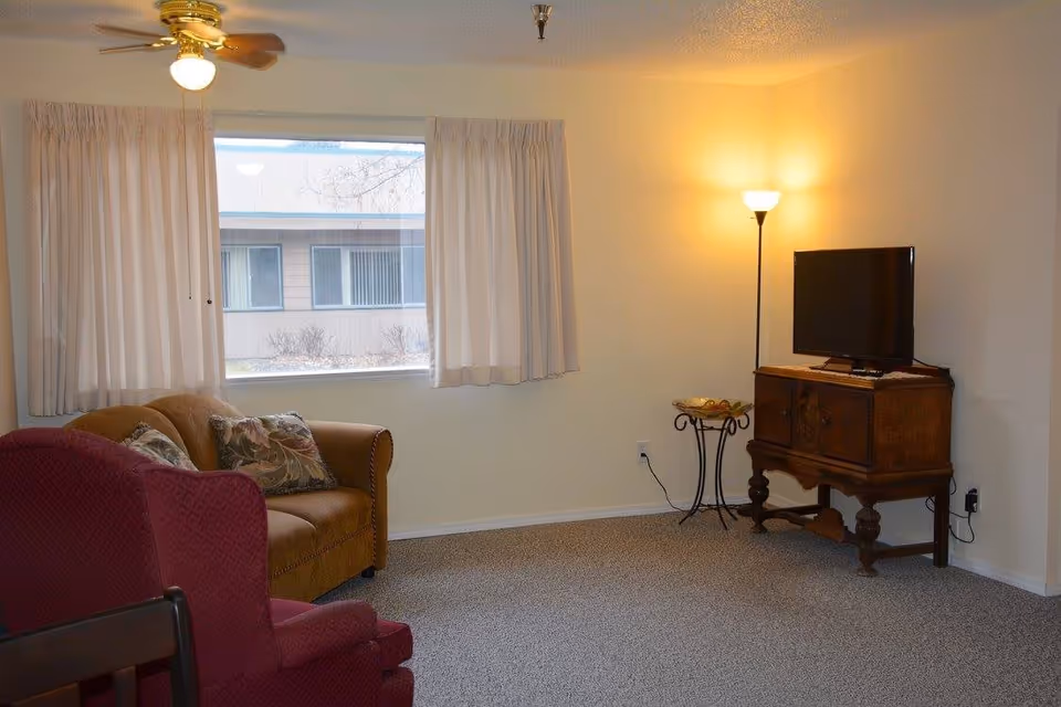 A cozy living room with a brown loveseat adorned with floral pillows, a red armchair, a wooden cabinet with a flat-screen TV on top, a floor lamp emitting warm light, and a window with white curtains showing an exterior building view.