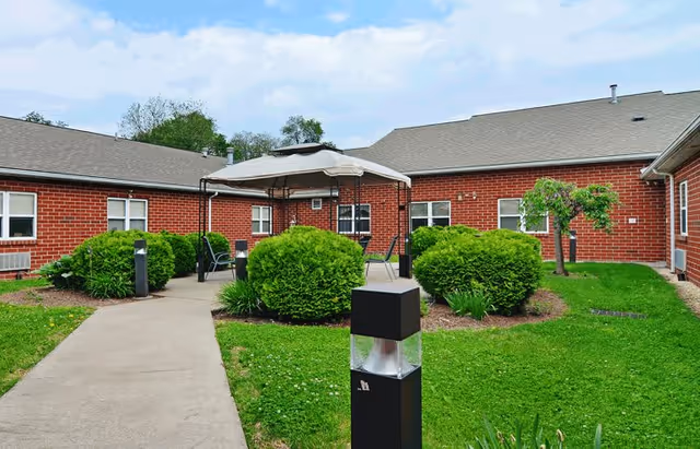 Outdoor courtyard area of a senior living facility with a concrete walkway, green bushes, a canopy with chairs underneath, and red brick buildings surrounding the space under a partly cloudy sky.