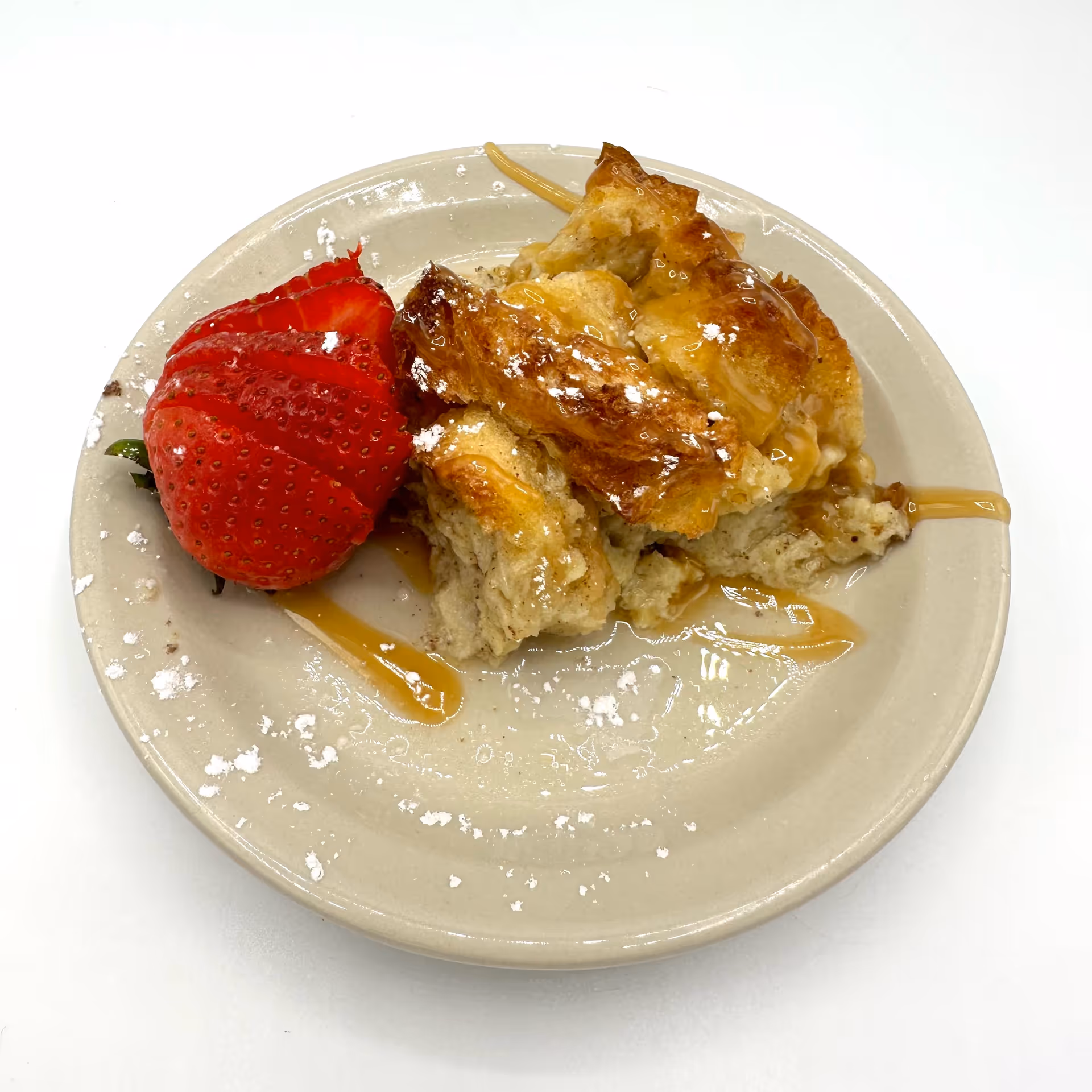A beige plate with a serving of bread pudding drizzled with caramel sauce and dusted with powdered sugar, accompanied by a sliced strawberry.