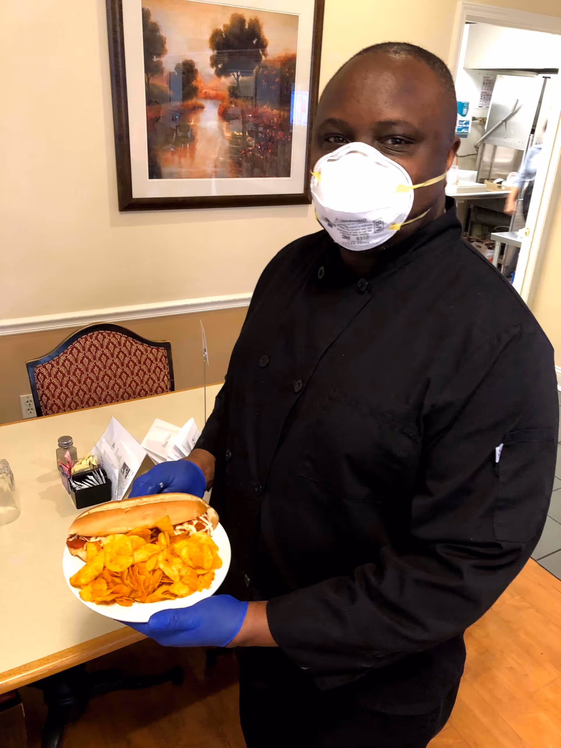 A person wearing a black chef's coat, blue gloves, and a white face mask is holding a plate with a sandwich and potato chips in a dining area. Behind them is a table with a red patterned chair, a condiment holder, and a framed painting on the wall. A kitchen area is visible through an open door in the background.