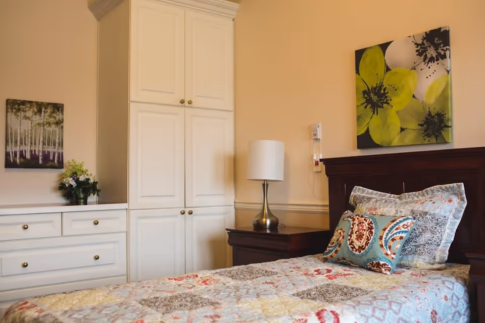 A bedroom with a single bed featuring a colorful quilt and decorative pillows. Next to the bed is a dark wooden nightstand with a silver lamp. The wall behind the bed has a large painting of green and white flowers. On the left side of the room, there is a white dresser with drawers and a tall white cabinet. A small vase with flowers and a framed picture of trees are placed on the dresser.