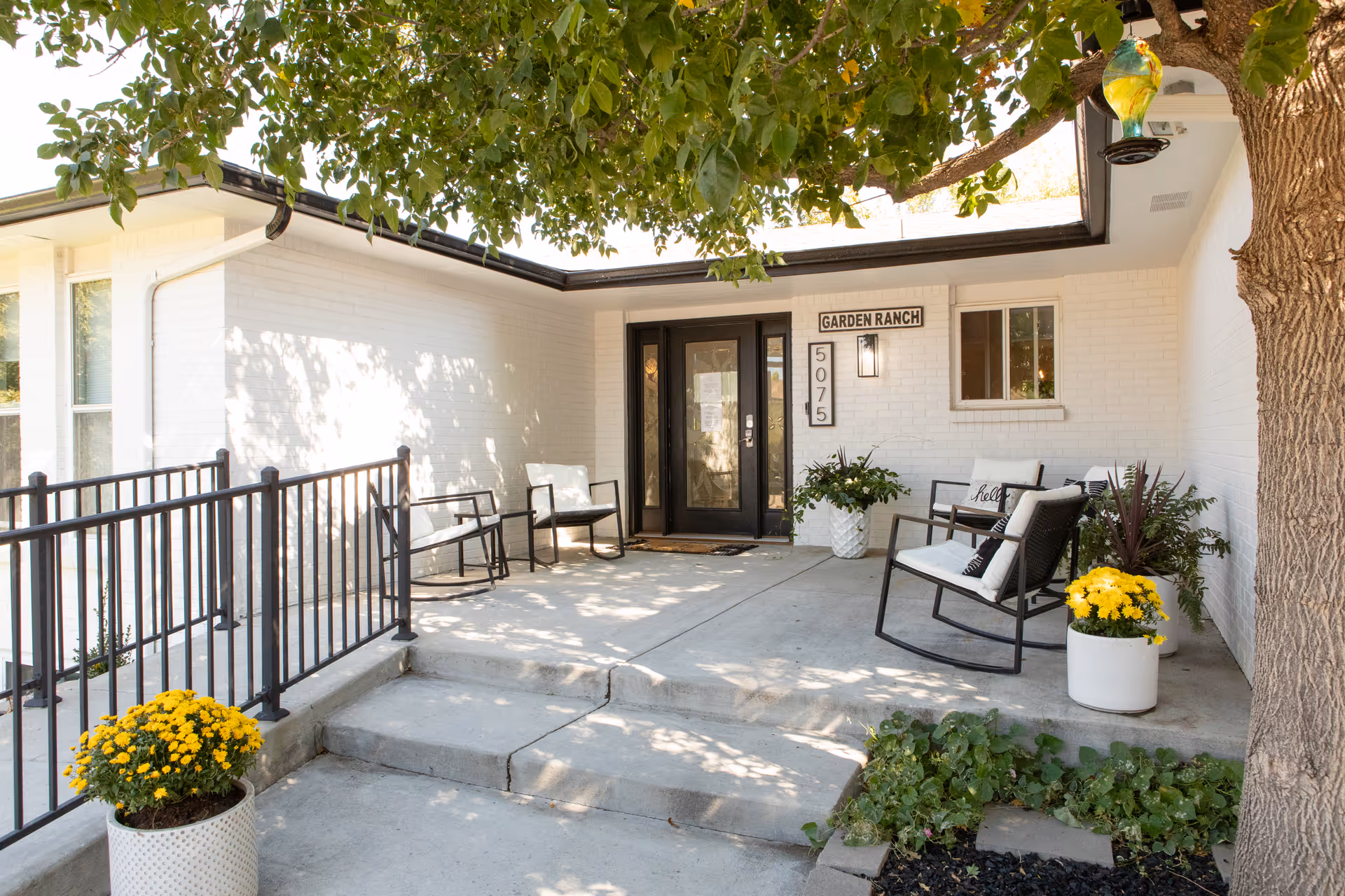 Front entrance of Garden Ranch Assisted Living featuring a white brick exterior, black double doors with glass panels, a sign with the facility name and address number 5075, a concrete porch with steps and a black metal railing, several white cushioned rocking chairs, potted yellow flowers, and a tree providing shade.