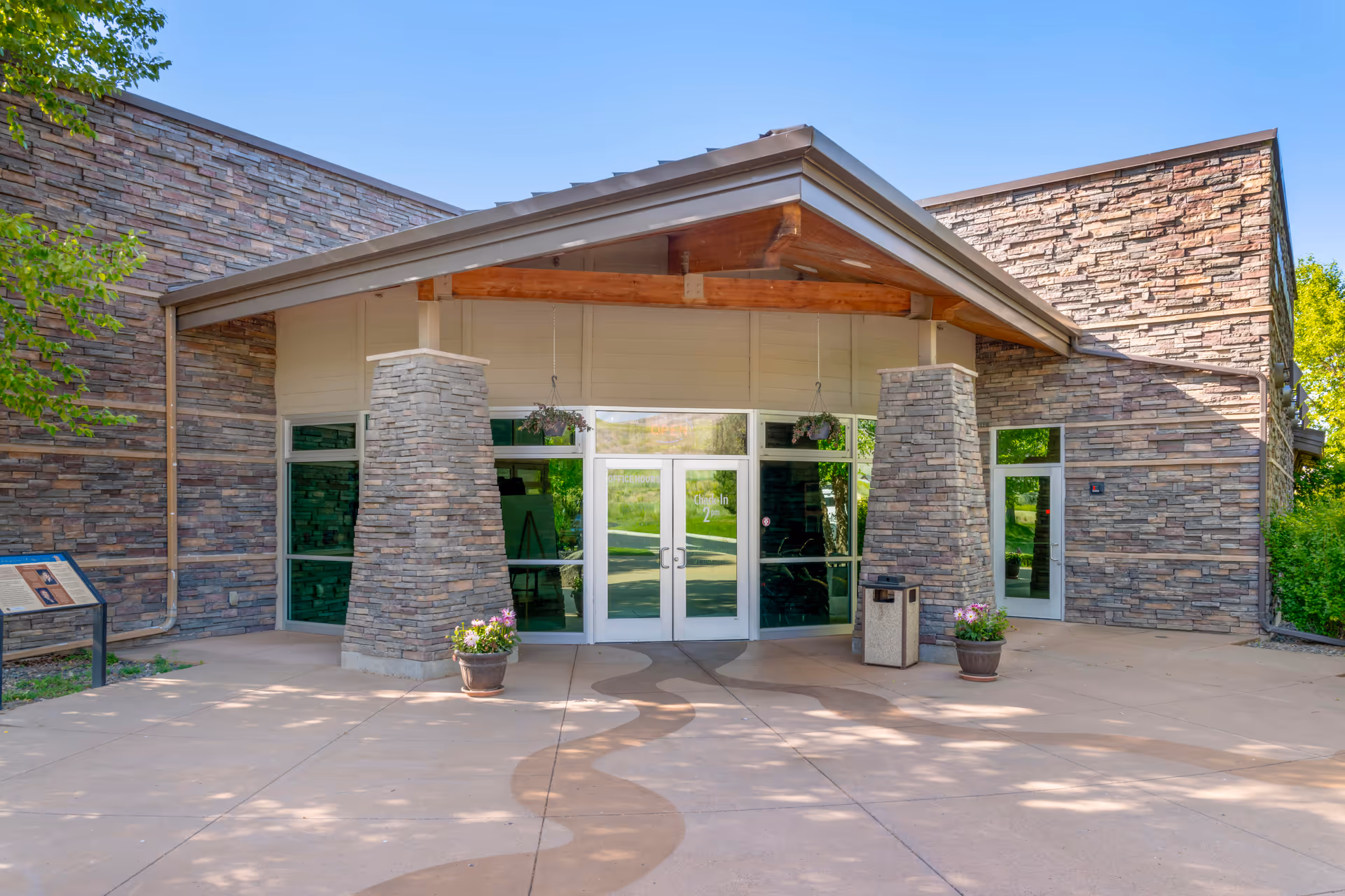 Entrance to a building with stone walls and large glass double doors under a peaked roof supported by two stone pillars. There are potted plants on either side of the entrance and a trash bin to the right. The area is well-lit with natural sunlight and surrounded by greenery.