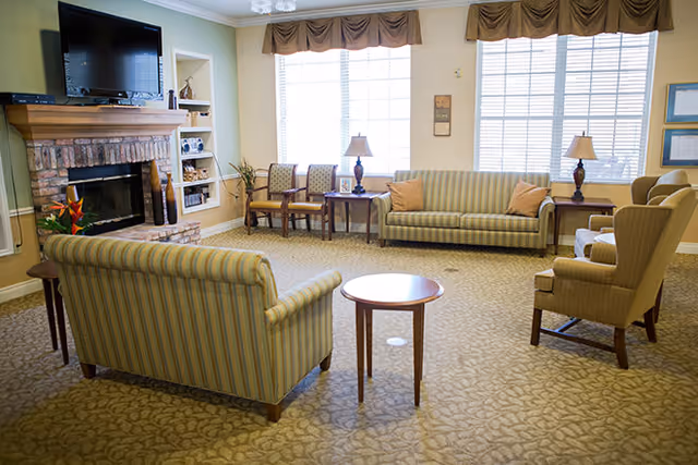 Bright communal living room featuring striped sofas and armchairs arranged around a fireplace and TV beneath large windows.
