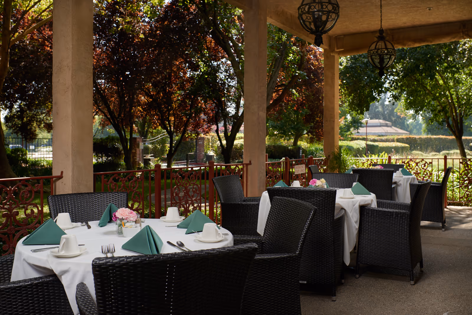 Outdoor covered patio dining area with several tables set with white tablecloths, green folded napkins, white cups and saucers, and small flower arrangements. The patio is surrounded by a decorative red railing and shaded by large trees with green and reddish leaves.