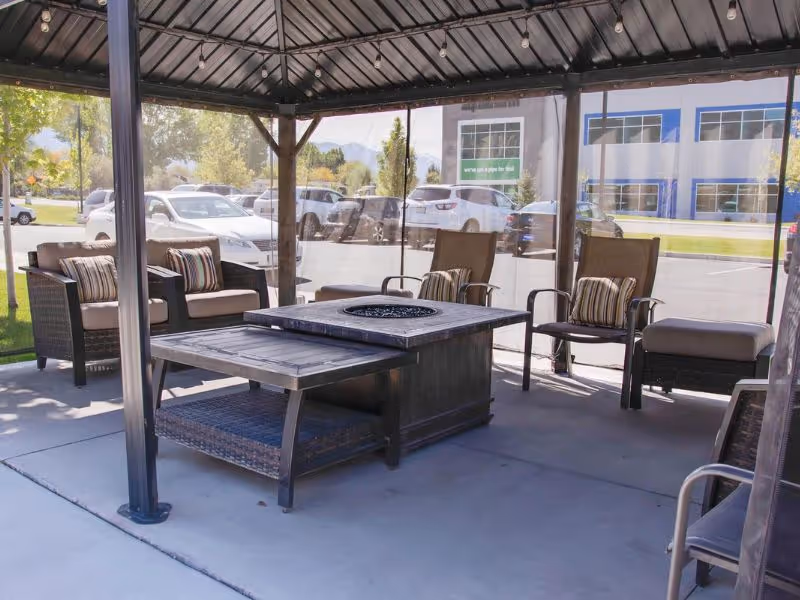 Outdoor seating area under a gazebo with cushioned chairs and a central fire pit table, located near a parking lot with cars and a building in the background.