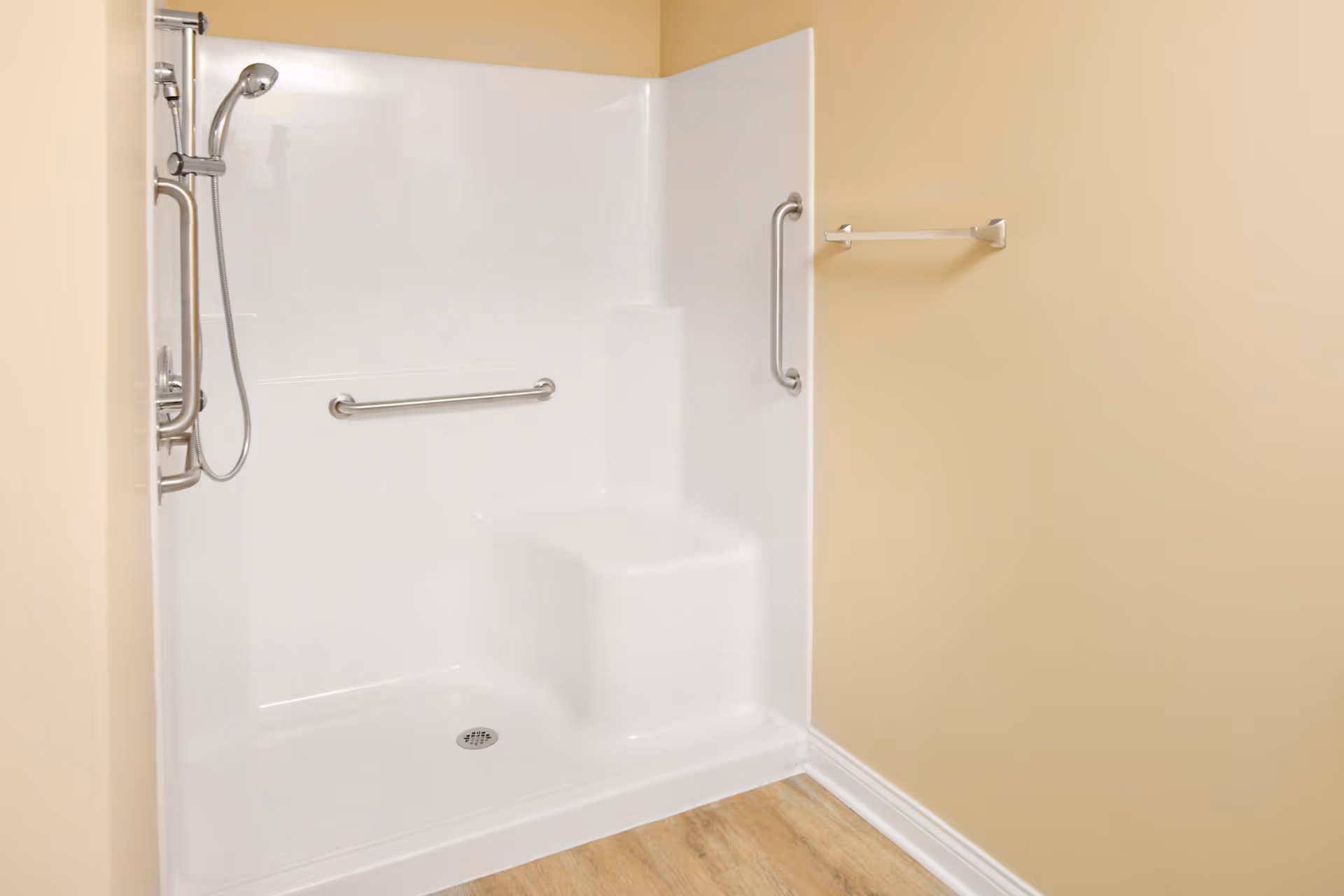A clean, accessible shower area with beige walls and wood-look flooring. The shower has a built-in seat, stainless steel grab bars, and a handheld showerhead. There is also a towel rack mounted on the wall outside the shower.