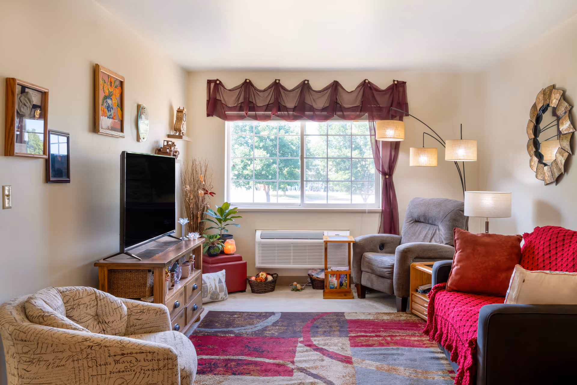 A cozy living room with a large window covered by sheer purple curtains. The room features a patterned armchair, a wooden TV stand with a flat-screen TV, a gray recliner chair, and a dark sofa with red and beige cushions and a red throw blanket. There is a colorful area rug on the floor, a decorative wall mirror, and several framed pictures on the walls. A floor lamp with three lights and a table lamp provide lighting.