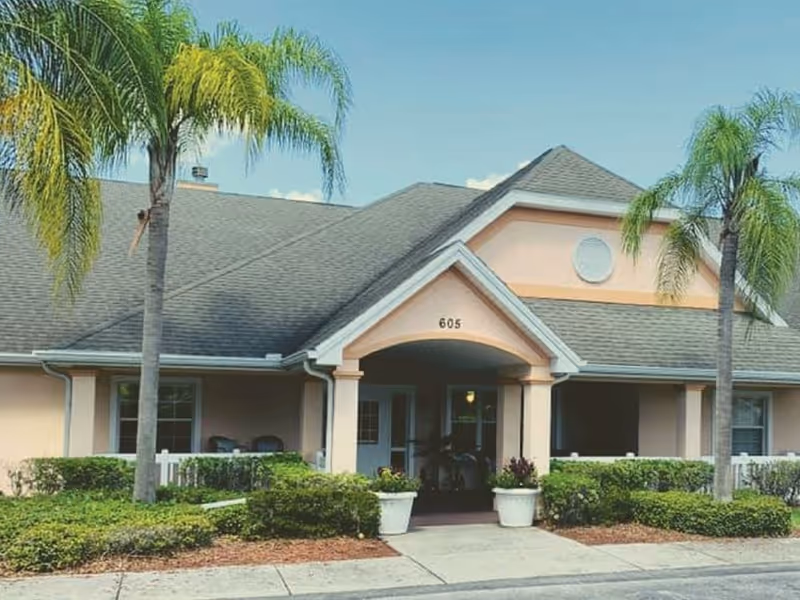 Exterior view of a single-story building with a gray shingled roof and peach-colored walls, featuring a covered entrance with the number 605 above it. The building is surrounded by green shrubs and palm trees under a clear blue sky.