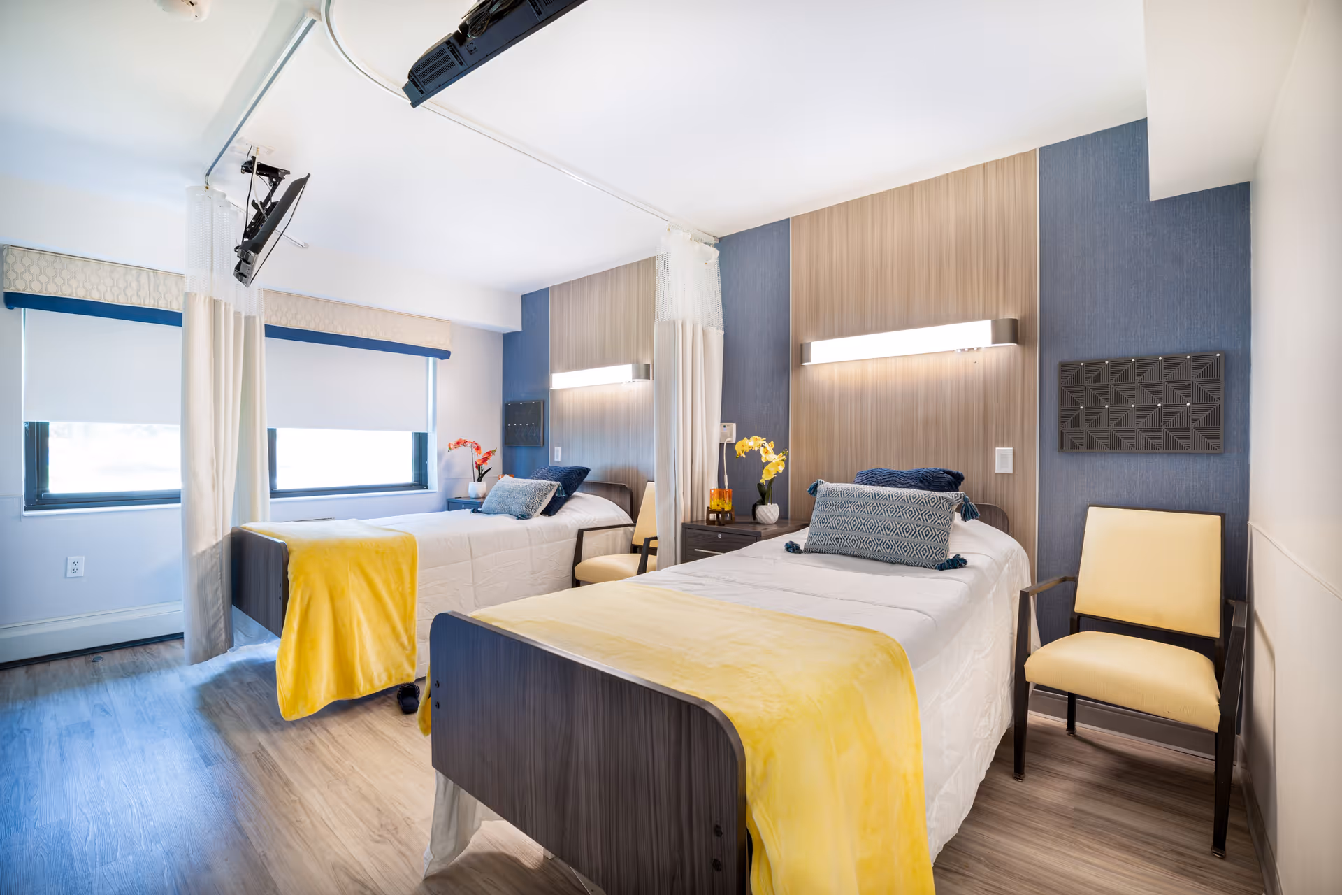 A bright and clean nursing facility bedroom with two single beds, each with white bedding and yellow throws. The room features wood flooring, blue accent walls behind the beds, wall-mounted lights, a chair beside each bed, and two windows with white roller shades. There are also ceiling-mounted televisions above each bed and privacy curtains between the beds.