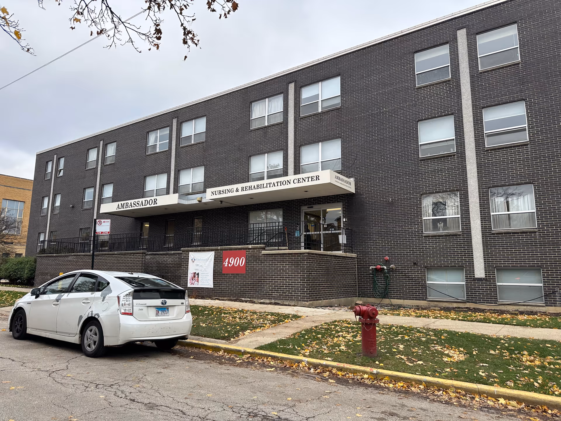 Exterior view of a three-story dark brick building with multiple windows and a white awning that reads 'Ambassador Nursing & Rehabilitation Center'. A white car is parked on the street in front of the building, and a red fire hydrant is visible on the grassy area near the sidewalk. The sky is overcast and there are some fallen leaves on the ground.
