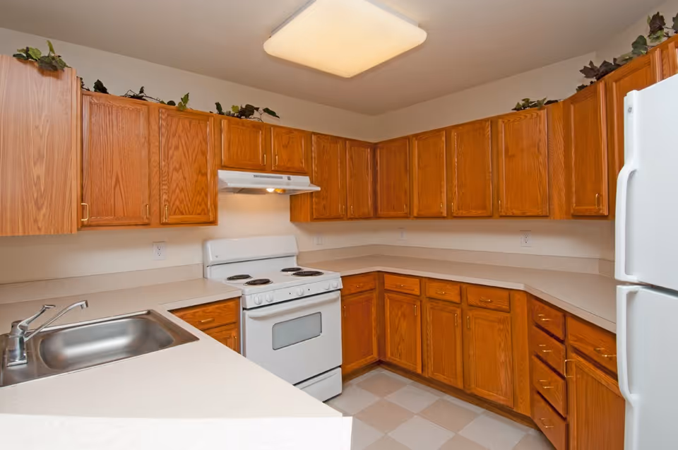 A kitchen with wooden cabinets, a white electric stove with a range hood, a white refrigerator, a stainless steel sink, and beige countertops. The floor has a checkered pattern with beige and white tiles. There are decorative green plants placed on top of the cabinets.