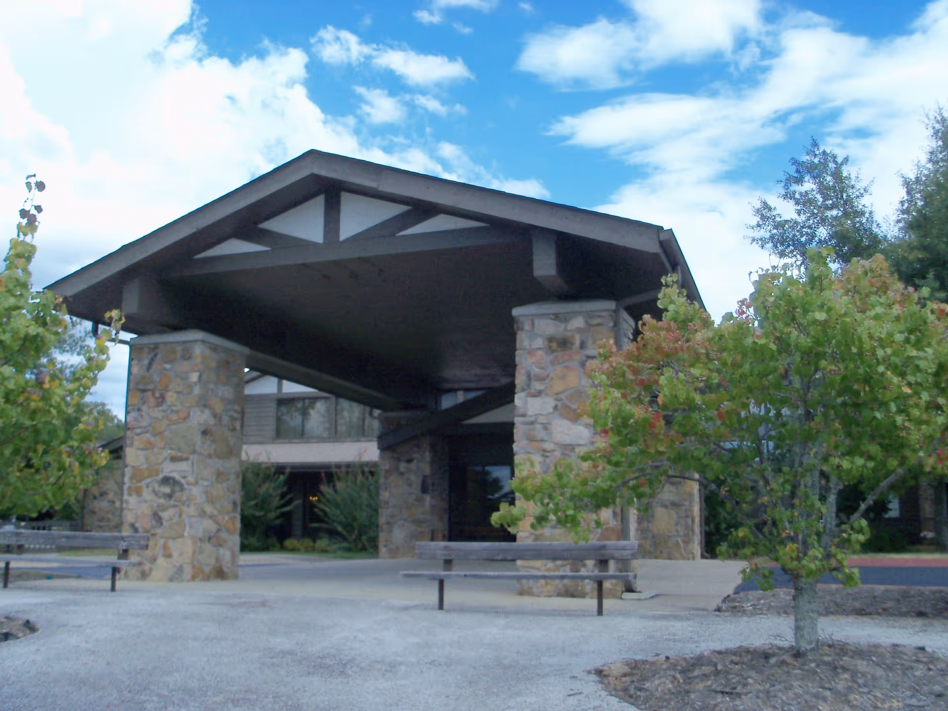 Exterior view of a building entrance with a large covered porch supported by stone pillars. There are benches on either side of the entrance and small trees with green and reddish leaves in the foreground. The sky is partly cloudy.