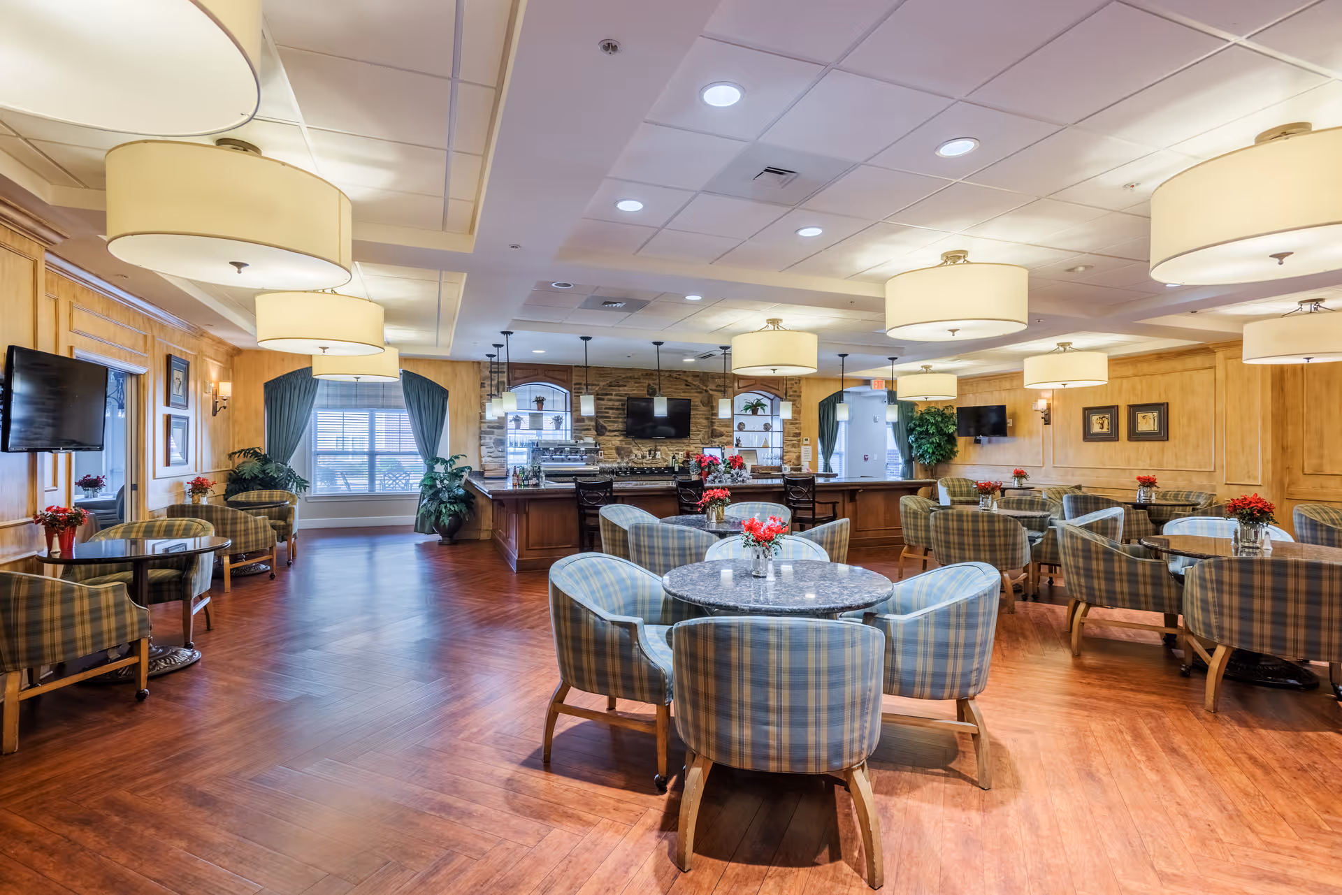 A spacious and well-lit dining room with multiple round tables surrounded by plaid upholstered chairs. The room features wooden flooring, large ceiling lights, wall-mounted televisions, and windows with green curtains. There are decorative plants and flower arrangements on the tables, and a serving area with a stone backsplash and shelves in the background.