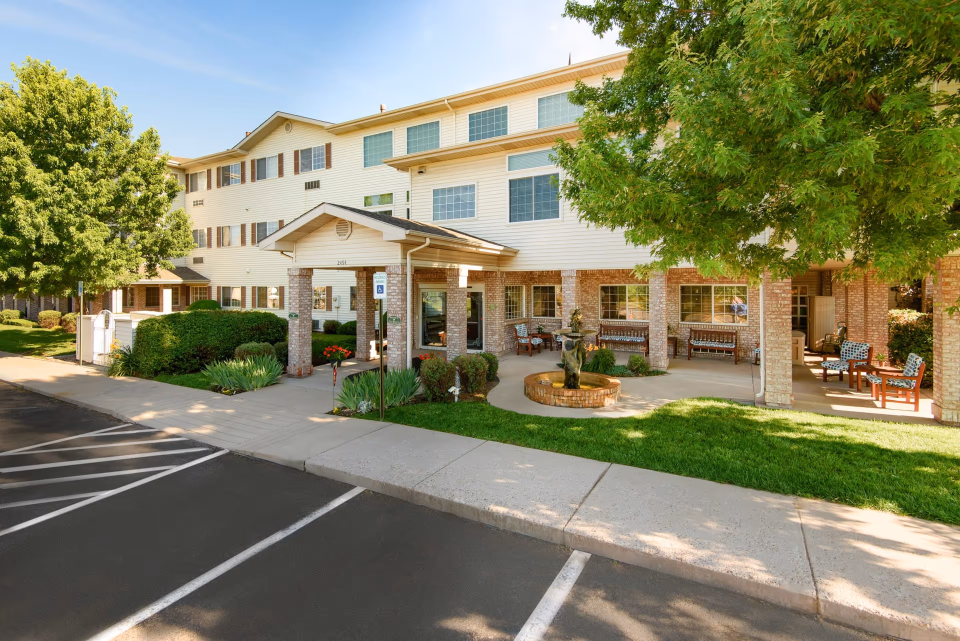 Exterior view of The Bridge At Colorado Springs senior living facility showing a three-story building with a covered entrance, brick pillars, outdoor seating area with chairs and benches, green lawn, trees, and a small fountain in front.