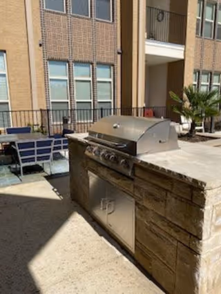Outdoor patio area with a built-in stainless steel grill and stone countertop. There are tables and chairs nearby, and the background shows a multi-story brick building with windows and balconies.