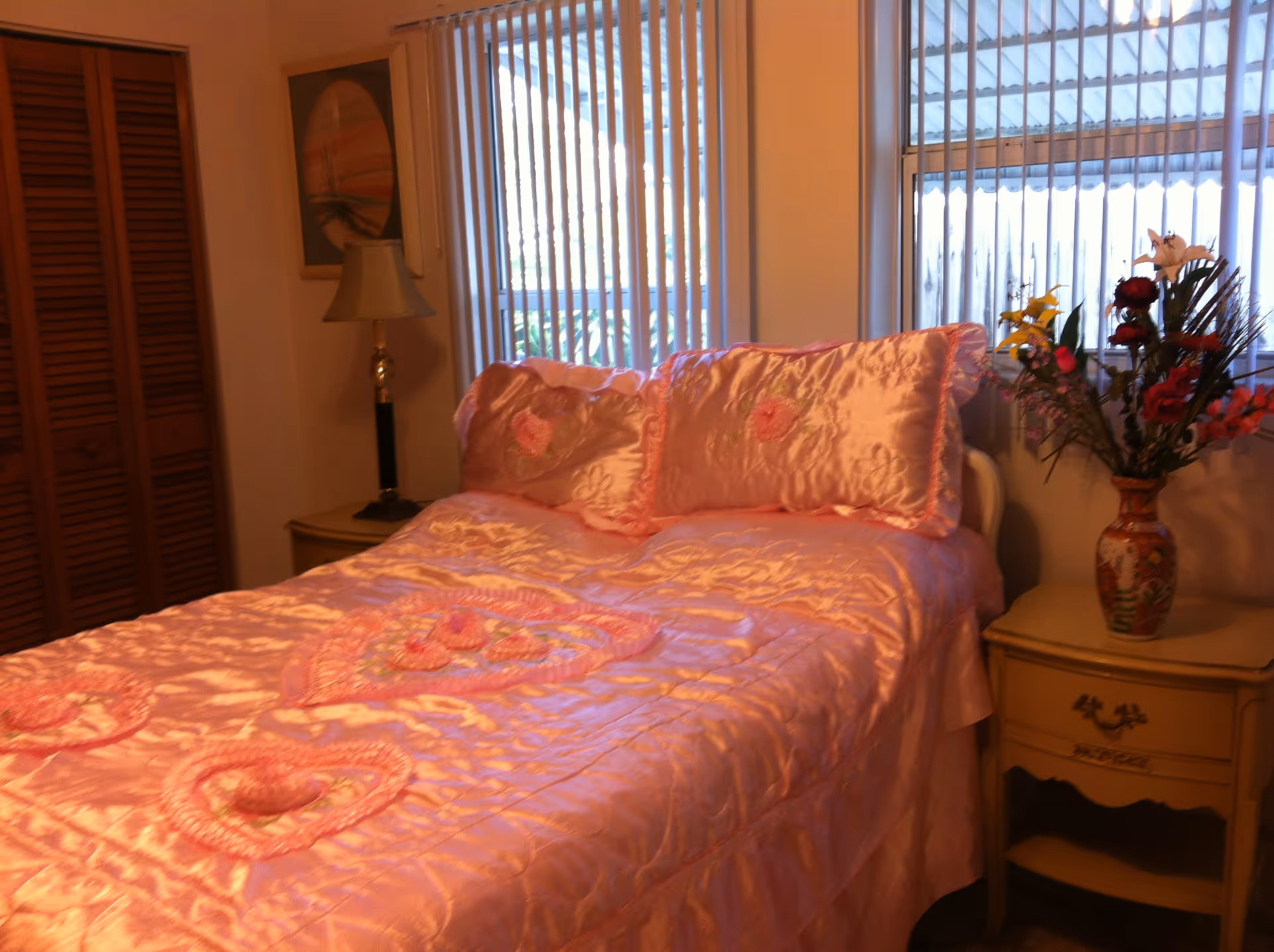 A bedroom with a bed covered in a shiny pink bedspread and matching pillows with heart designs. There is a nightstand with a floral vase and flowers on the right side of the bed, and a lamp on a small table on the left side. Vertical blinds cover the windows behind the bed.