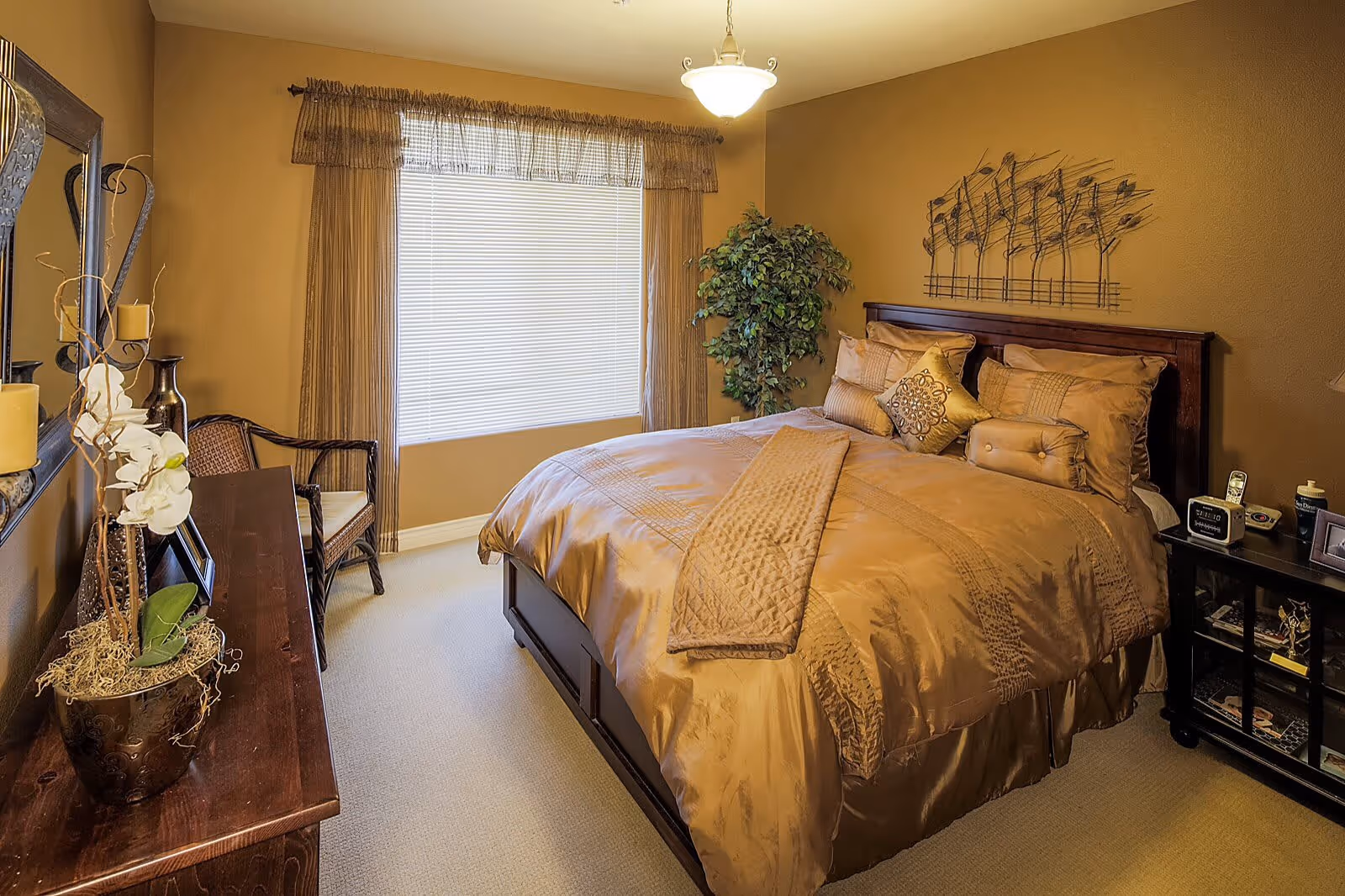 A cozy bedroom with a large bed covered in gold bedding and multiple pillows. There is a window with blinds and sheer curtains, a wooden dresser with decorative items including a potted orchid, a chair, a nightstand with a clock and phone, and a green potted plant in the corner. The walls are painted a warm golden color and there is a metal wall art piece above the bed.