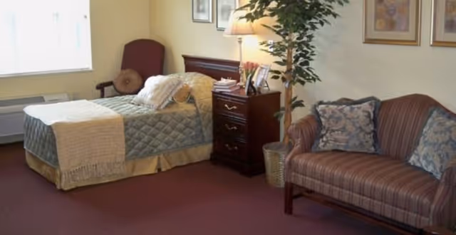 A furnished private room with a single bed, wooden nightstand and lamp, an armchair, potted plant, and a striped loveseat.