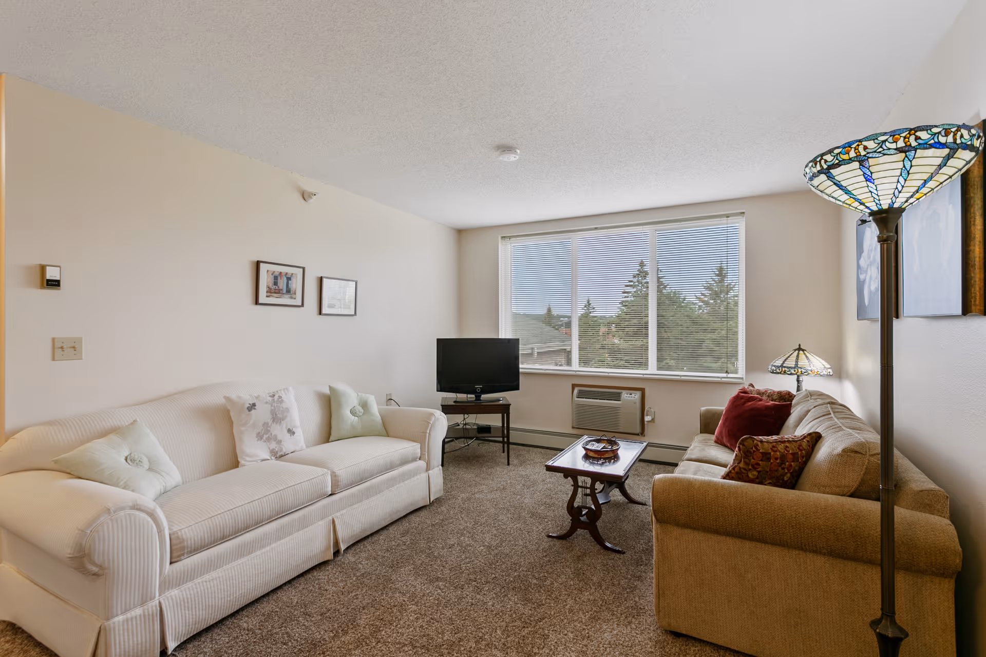 A cozy living room with two sofas facing each other, a small wooden coffee table in the center, a TV on a stand near a large window with blinds, and two decorative lamps with stained glass shades. The room has beige walls and carpeted floor.