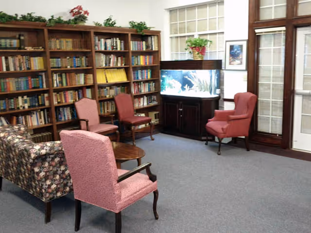 A cozy living room area with a floral patterned sofa, three pink upholstered chairs, a wooden coffee table, and a large bookshelf filled with books. There is a fish tank on a wooden cabinet near a window with blinds, and a potted plant on top of the cabinet. The room has carpeted flooring and a glass door with wooden framing.