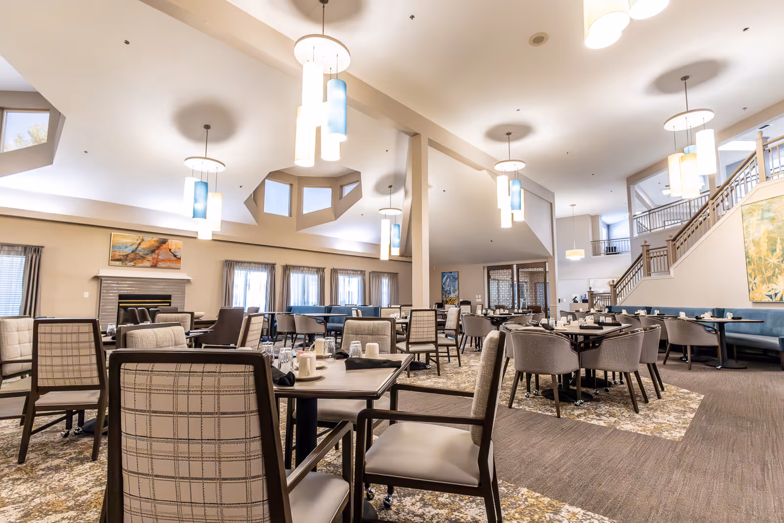 A spacious and well-lit dining room with multiple tables and chairs arranged neatly. The room features modern hanging light fixtures, large windows with curtains, a fireplace with artwork above it, and a staircase leading to an upper level. The decor includes neutral tones and patterned carpeting.