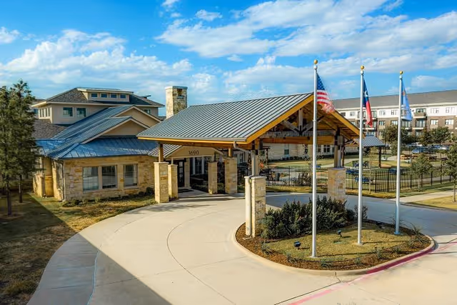 Entrance of The Auberge at Plano senior living facility featuring a covered driveway with stone pillars, three flagpoles with American, Texas, and another flag, and a multi-story building in the background under a partly cloudy sky.