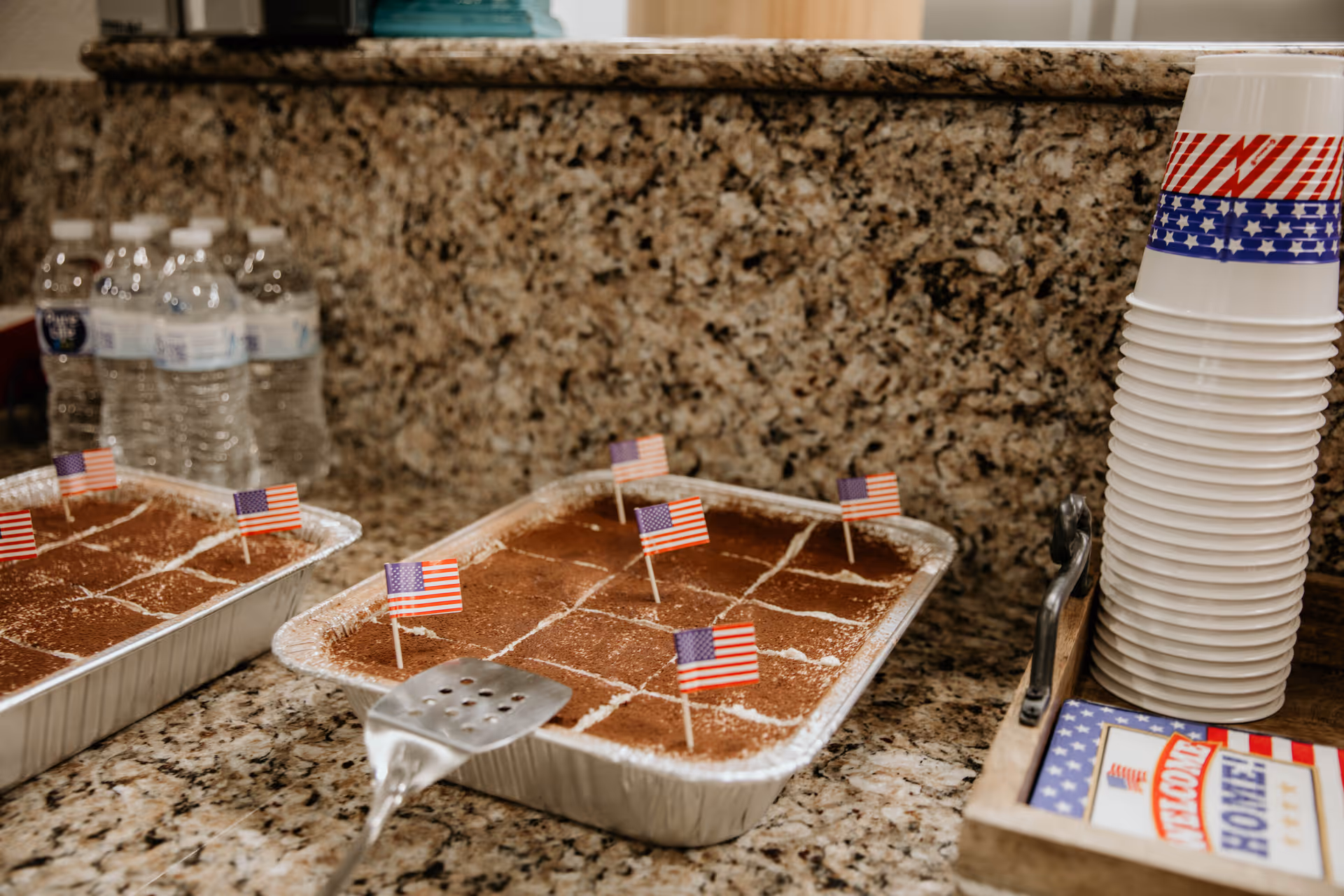 Two trays of dessert topped with small American flags on a granite countertop, with bottled water in the background and a stack of patriotic-themed disposable cups and napkins on the side.