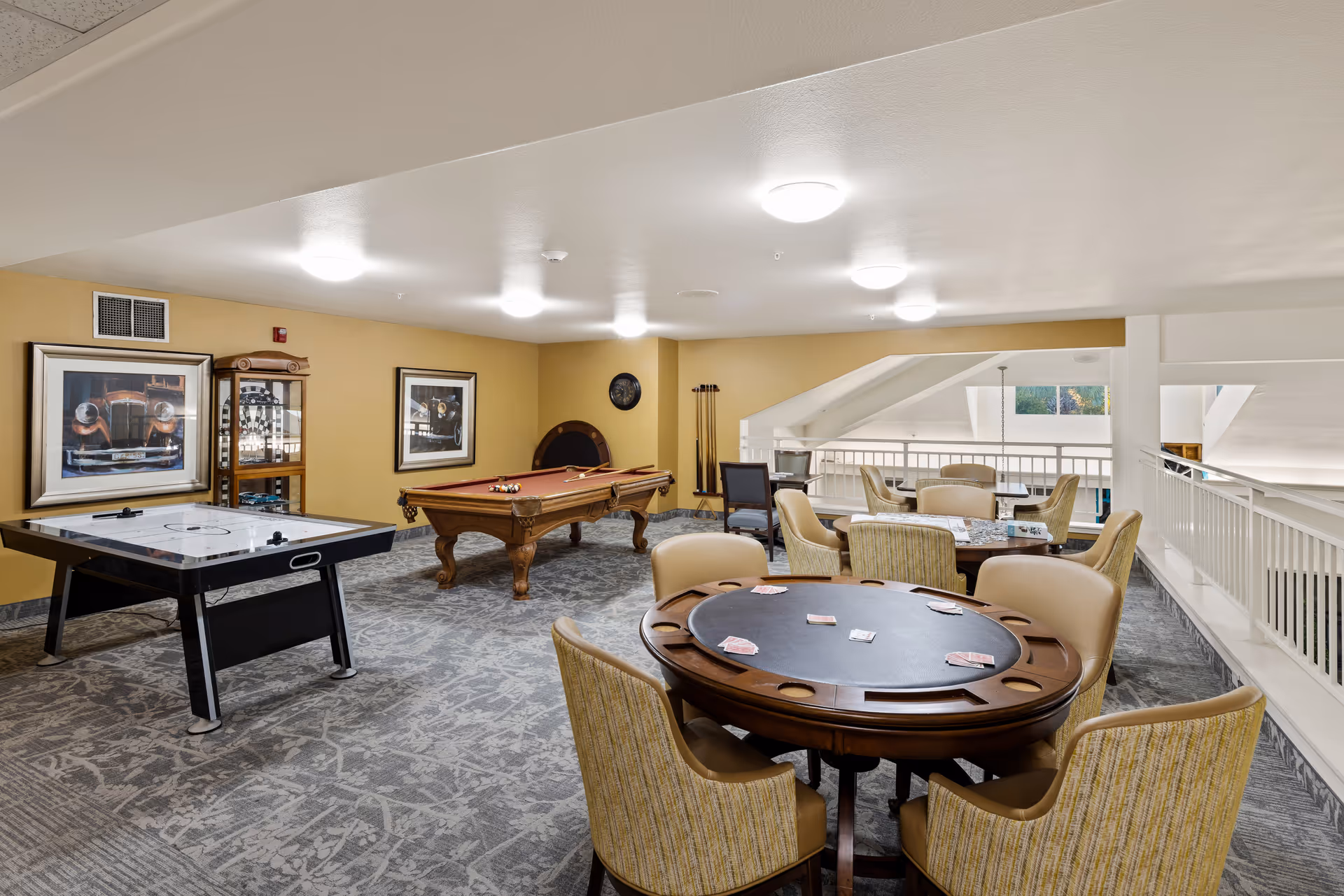 A senior living community game room featuring a pool table, air hockey table, and a round card table with chairs. The room has beige walls with framed pictures, carpeted floor, and ceiling lights. There is a railing overlooking a lower level and additional seating in the background.