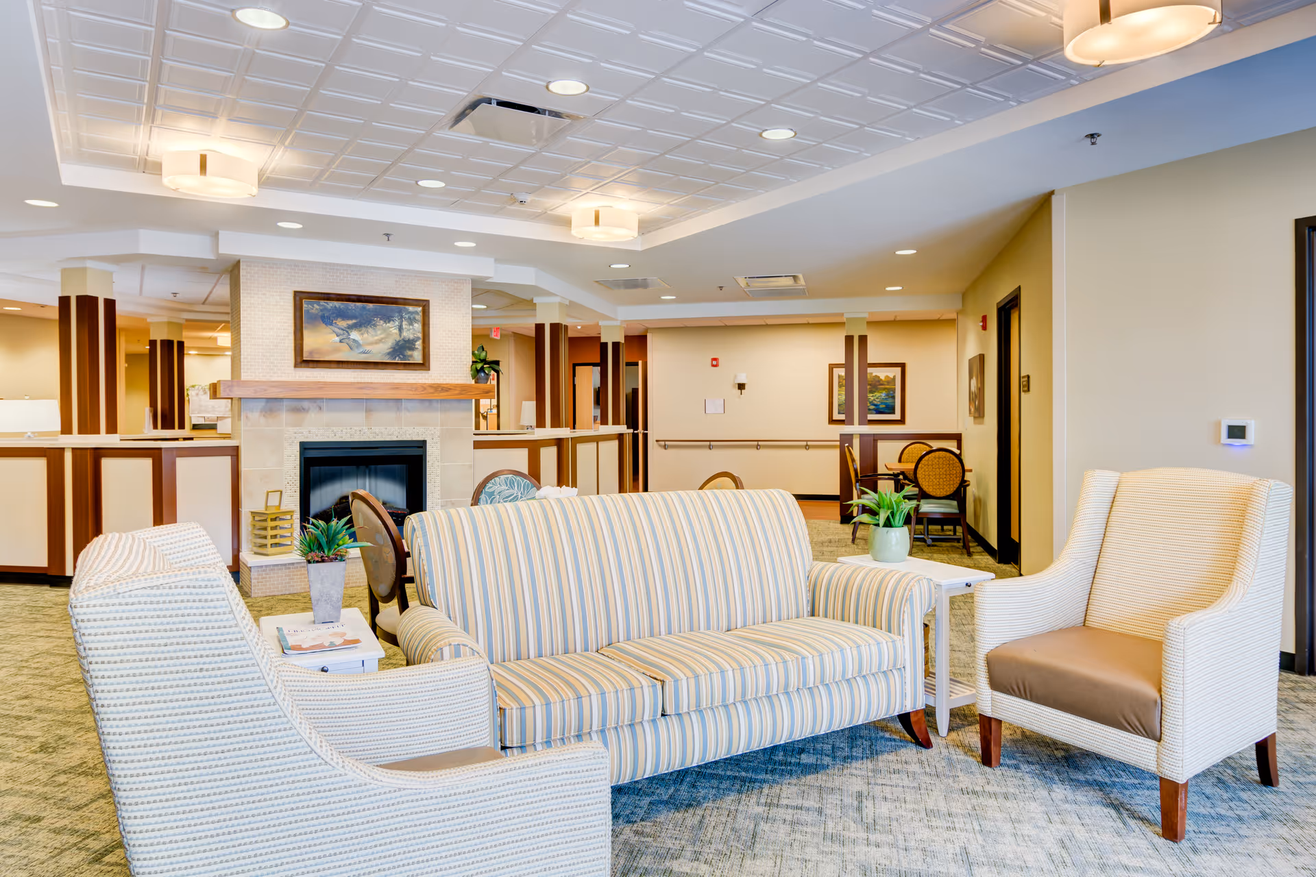 A bright and spacious living room area in a senior living facility with a striped sofa, two armchairs, a fireplace with a painting above it, and several small tables with potted plants. The room has a patterned carpet, recessed lighting, and a drop ceiling with square tiles.