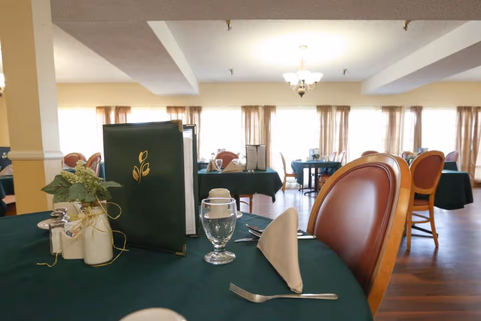 Dining room with tables covered in green tablecloths, set with menus, glassware, silverware, and napkins. Chairs with wooden frames and brown cushions surround the tables. Large windows with sheer curtains allow natural light to fill the room.
