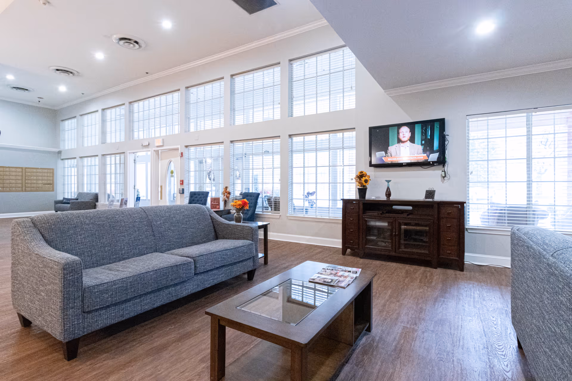 A bright and spacious living room area with large windows covered by white blinds, two gray upholstered sofas, a wooden coffee table with magazines, and a wooden TV stand with a flat-screen television mounted above it. The room has wood flooring and some decorative items like flowers on the tables.