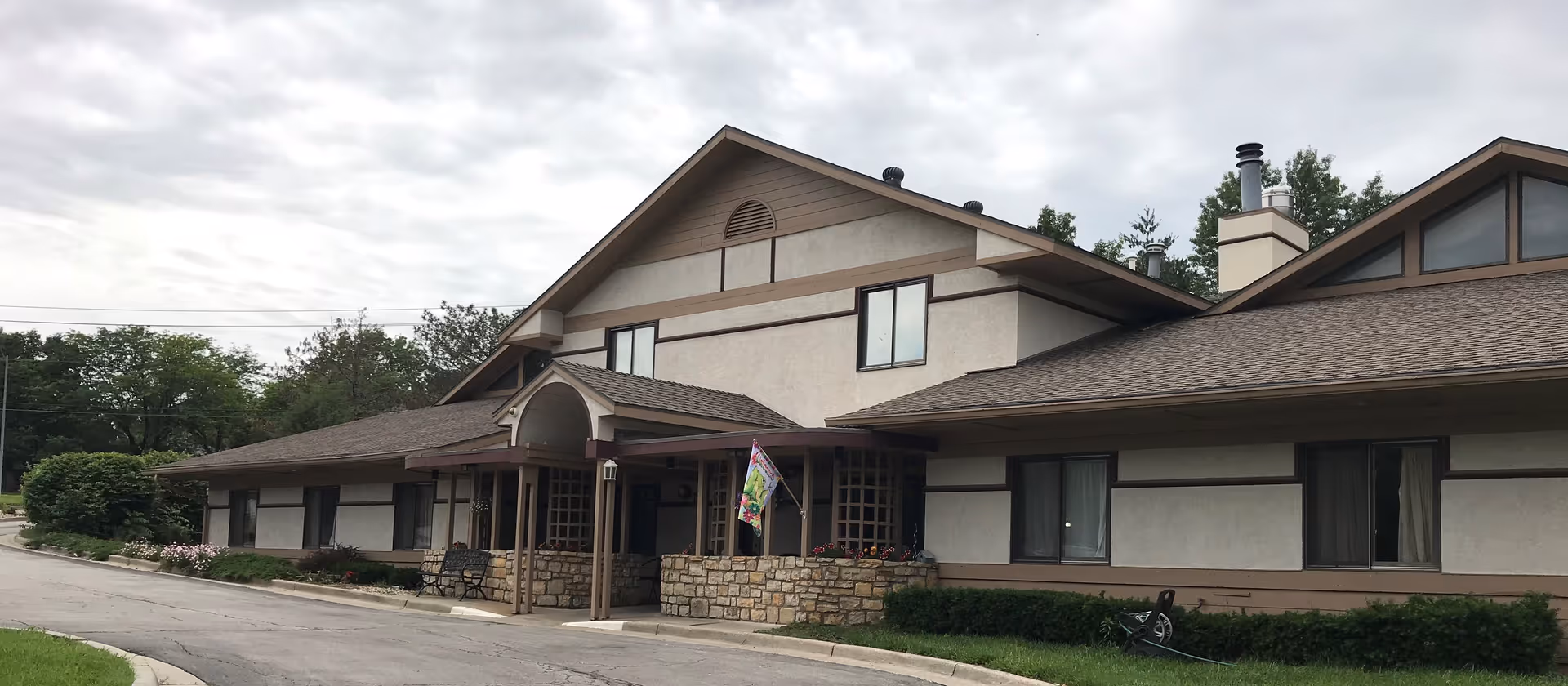 Front exterior of a two-story senior living building with a covered stone entry, driveway, and landscaping under a cloudy sky.