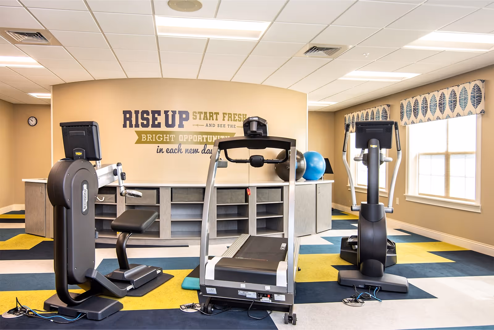 Bright fitness room with a treadmill, elliptical machines, exercise bike and an exercise ball beneath a 'RISE UP' motivational wall graphic.