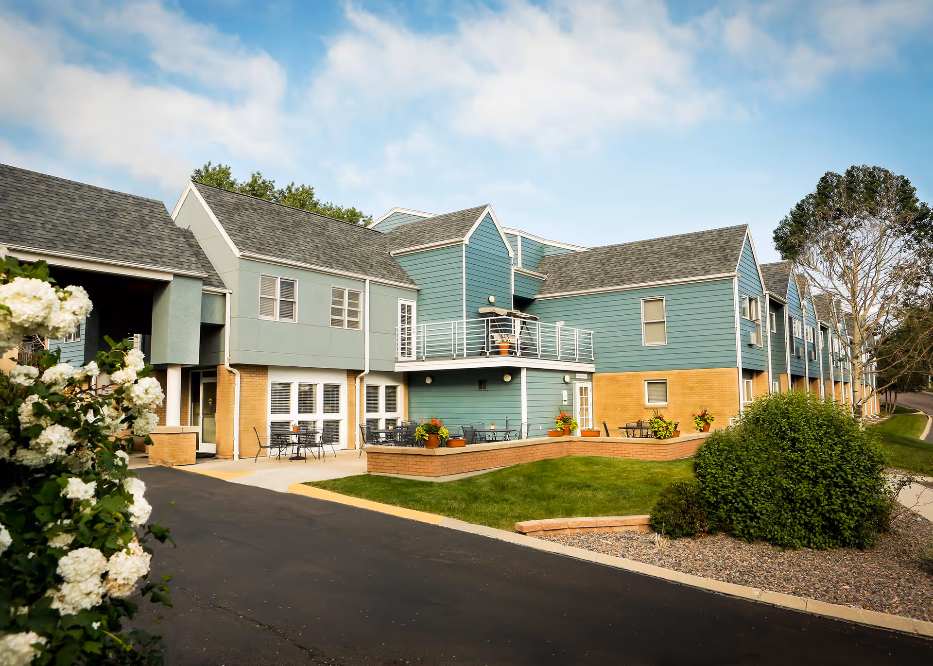 Exterior view of a two-story assisted living facility building with blue and beige siding, multiple windows, outdoor seating areas with tables and chairs, green lawn, bushes, and white flowers in the foreground under a partly cloudy sky.