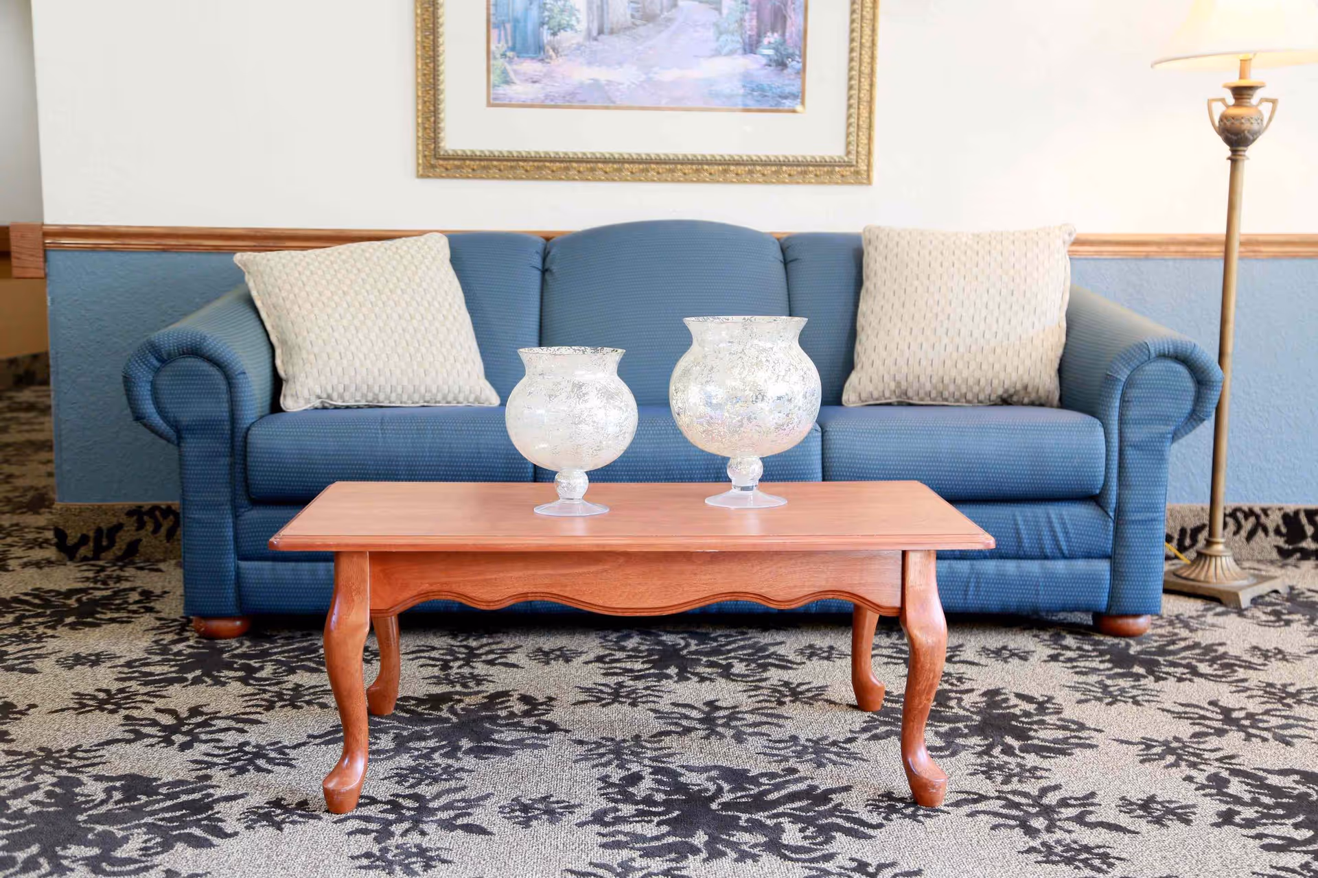A blue upholstered sofa with two beige patterned cushions is positioned against a wall with a framed painting above it. In front of the sofa is a wooden coffee table with two decorative glass candle holders. To the right of the sofa is a tall floor lamp with a beige lampshade. The floor is carpeted with a patterned design.