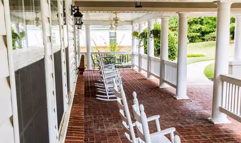 Covered brick porch with white rocking chairs and surrounding greenery.