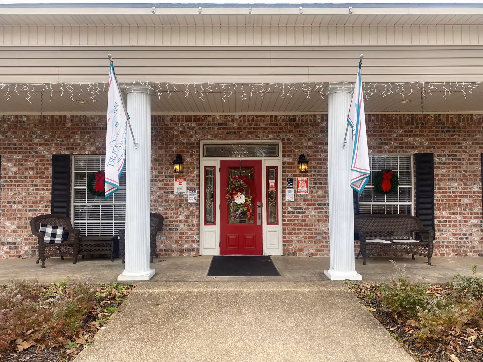 Front entrance of The Columns Of Bastrop facility featuring a red door with a decorative wreath, two white columns, brick exterior walls, two windows with black shutters and holiday wreaths, two wicker chairs with cushions on either side of the entrance, and two flags hanging from the columns.