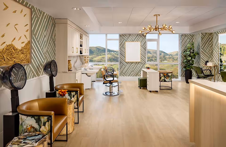 A bright and spacious hair salon area with large windows showing a scenic view of green hills. The room features salon chairs, hair dryers, a reception desk, patterned green and white wallpaper, and modern light fixtures. There are also comfortable seating areas with floral-patterned chairs and small wooden tables.