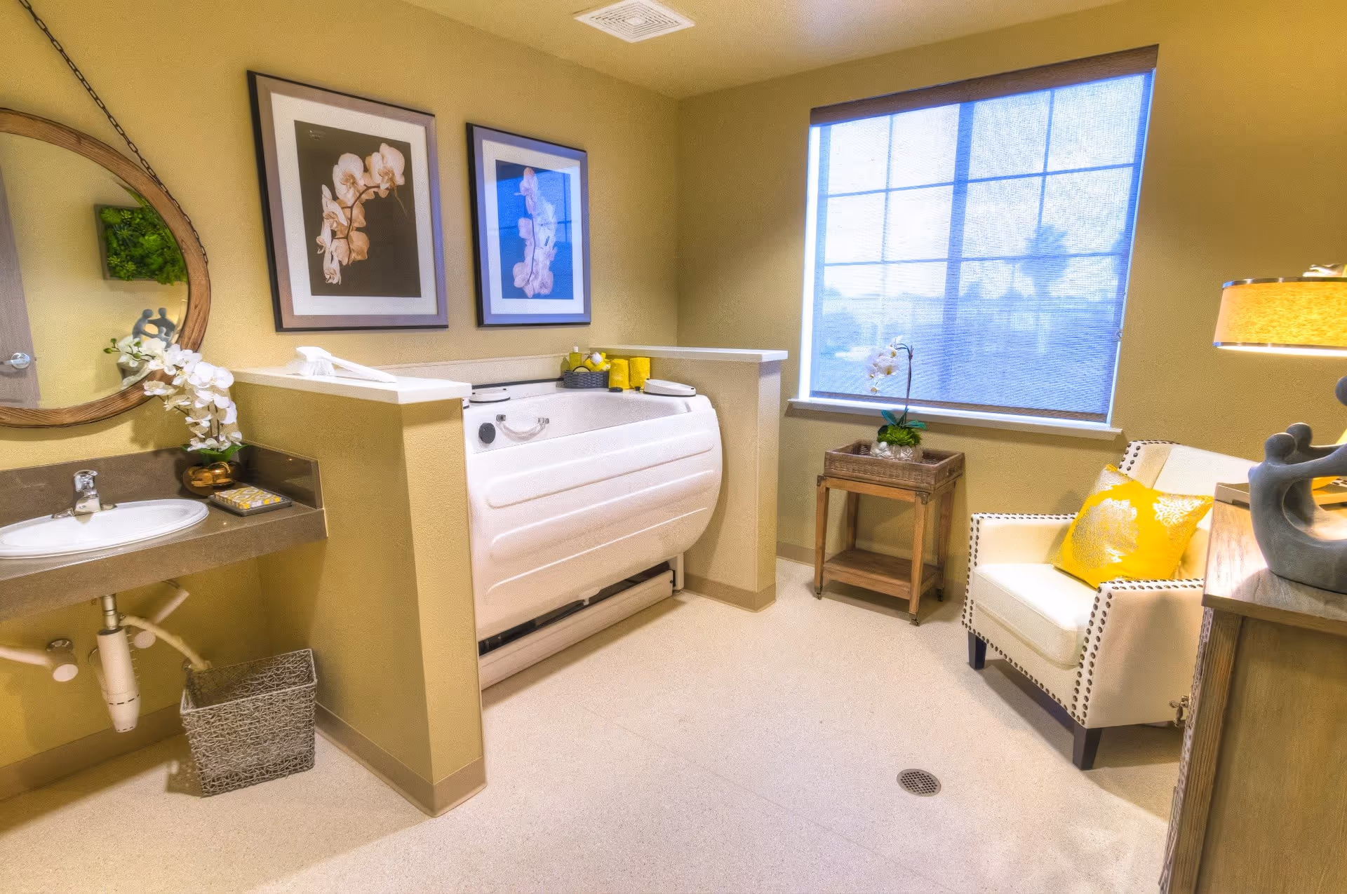 A bright and clean bathroom in a memory care facility featuring a white walk-in bathtub with safety handles, a sink with a round mirror above it, two framed floral pictures on the wall, a small wooden side table with a plant, a comfortable armchair with yellow cushions, and a window with a blue shade letting in natural light.
