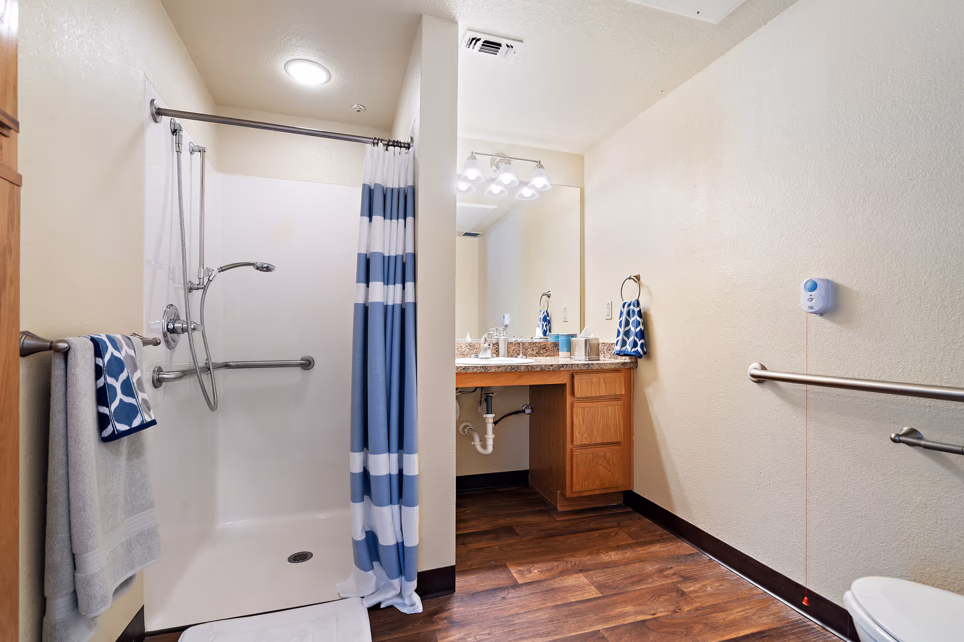 A bathroom with a walk-in shower featuring a blue and white striped shower curtain, grab bars, and a handheld showerhead. There is a wooden vanity with a granite countertop, a large mirror, and a light fixture above. The bathroom has wood-look flooring and beige walls, with towels hanging on the wall and a pull cord emergency alert system.