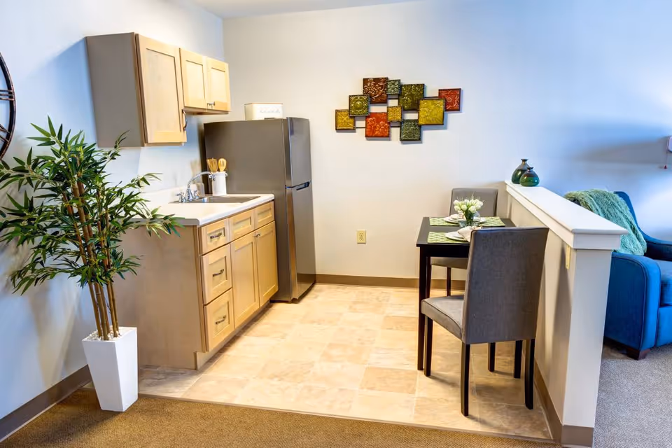 A small kitchen area with light wood cabinets, a stainless steel refrigerator, and a sink. Next to the kitchen is a small dining table set for two with green placemats and a flower centerpiece. A decorative wall art piece with colorful square panels hangs on the wall above the dining table. A potted plant is placed on the left side near the kitchen, and a blue armchair with a green throw blanket is partially visible on the right side.