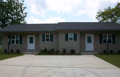 Single story senior living building with multiple entrances, green shutters, and a concrete driveway.