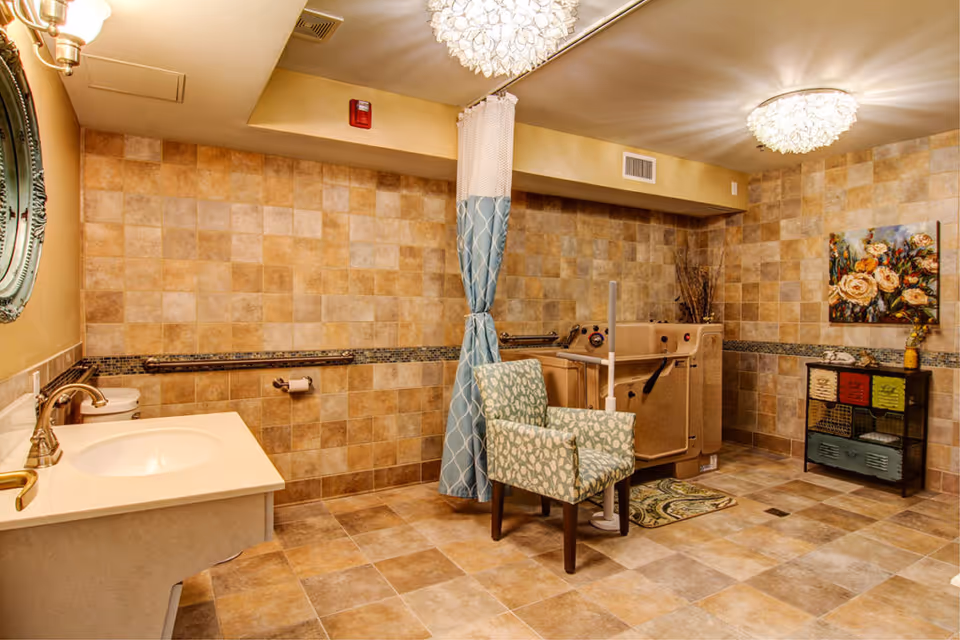 A spacious bathroom with beige and tan tiled walls and floor. The room features a white sink with a gold faucet, a toilet, a patterned armchair, a bathtub with safety rails, and a blue and white shower curtain. There is a decorative floral painting on the wall above a small cabinet with colorful drawers. The ceiling has two ornate light fixtures.