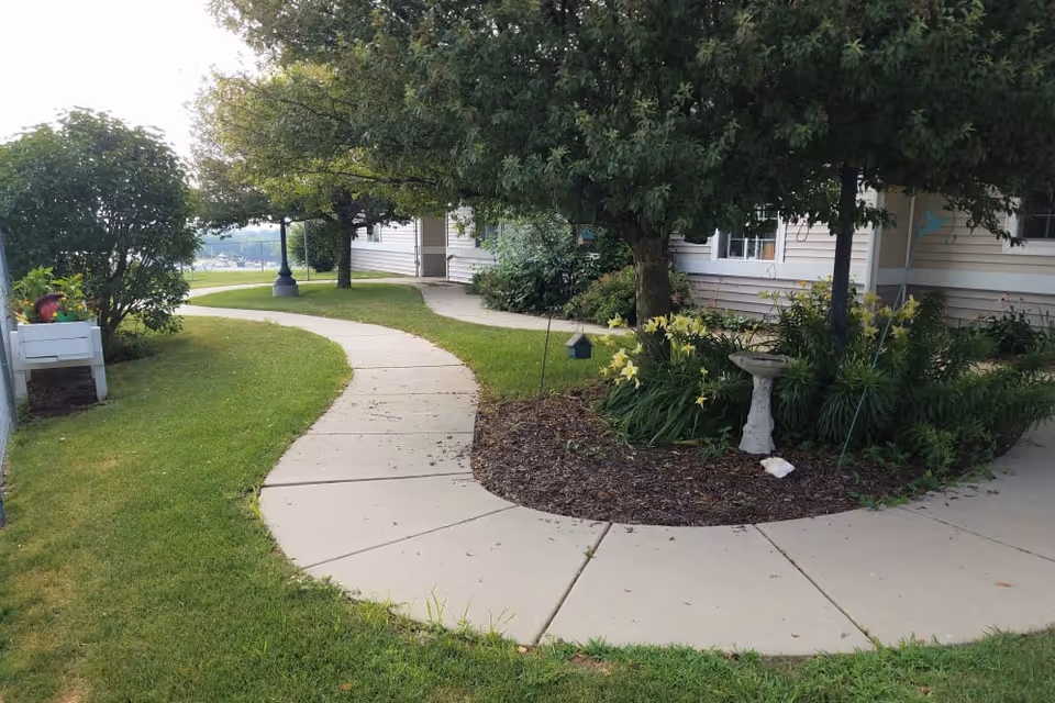 Curved concrete walkway surrounded by green grass, bushes, and trees leading to the entrance of a beige building with white trim. There is a birdbath and yellow flowers in a mulched garden bed near the walkway.