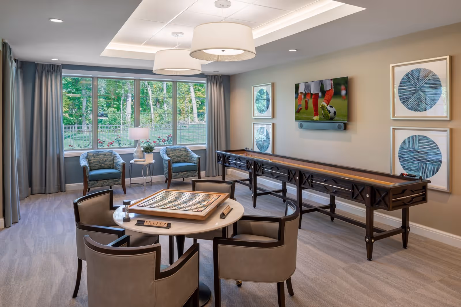 A bright and spacious game room with large windows showing a green outdoor view. The room features a round table with four chairs and a board game set up on it. There is a long shuffleboard table against the wall, a flat-screen TV mounted above it displaying a soccer game, and two armchairs with a small table and lamp between them near the windows. The walls are decorated with framed abstract art, and two large ceiling lights hang above the table.
