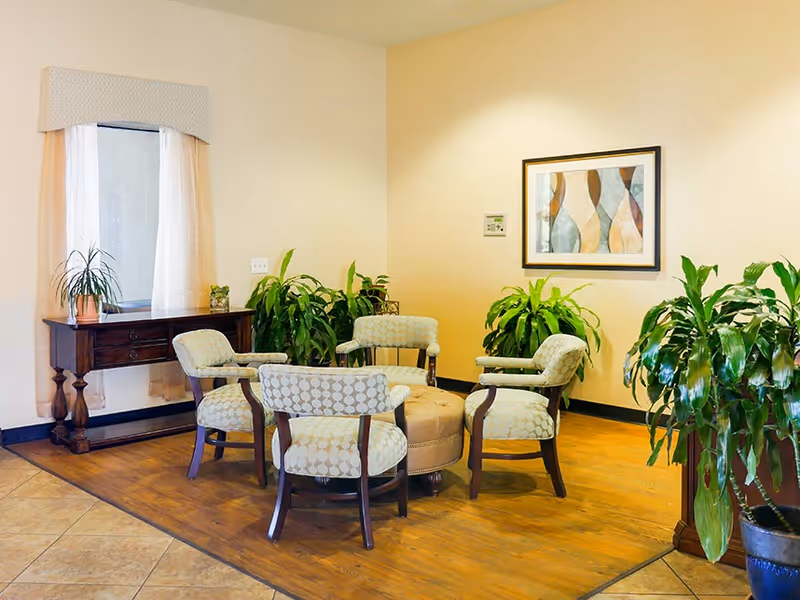 Small seating area with five upholstered chairs arranged around a central ottoman, potted plants, a side table under a window, and framed artwork on a light wall.