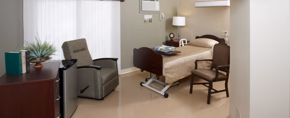 A nursing facility bedroom with a hospital-style bed covered in beige bedding, a brown wooden chair beside the bed, a gray recliner chair near a window with vertical blinds, a small wooden dresser with books and a plant on top, and a small refrigerator underneath the dresser. The room has beige walls and a beige floor.